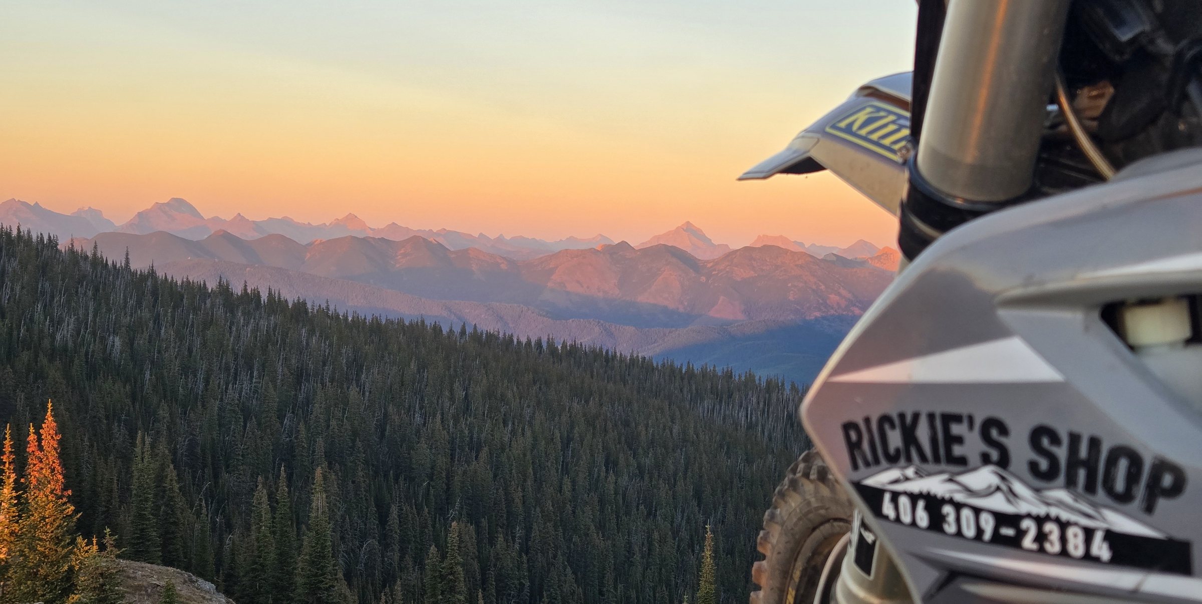 A dirt bike with a Rickie's Shop plate parked on a mountain ridge overlooking the Flathead Valley at sunset